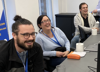 Three people sat at a desk laughing, there are two men and one woman.