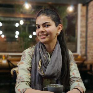 A woman with long brown hair, sat with a drink and wears a scarf.
