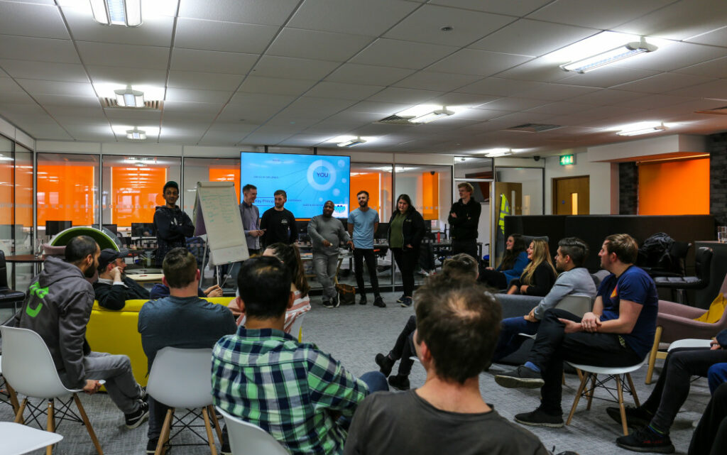 A group of people sit and listen to seven people talking about a subject.
