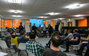 A group of people sit and listen to seven people talking about a subject.