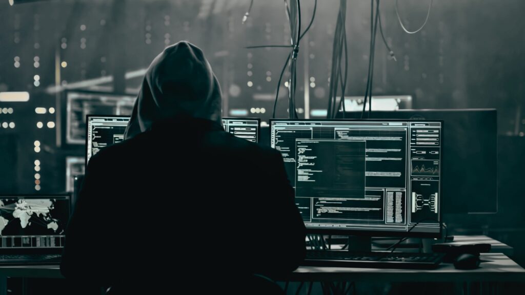 A hooded figure sits in front of multiple laptop screens.