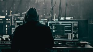 A hooded figure sits in front of multiple laptop screens.