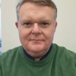 A man with shirt silver hair wears a green jumper.